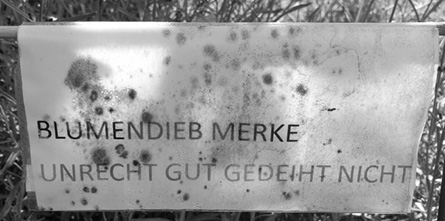A weathered sign with spots and stains reads "BLUMENDIEB MERKE UNRECHT GUT GEDEIHT NICHT" in German.
Description generated by AI