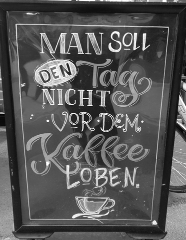 A chalkboard sign with decorative lettering in German reads "Man soll den Tag nicht vor dem Kaffee loben" with a drawing of a steaming coffee cup at the bottom.
Description generated by AI