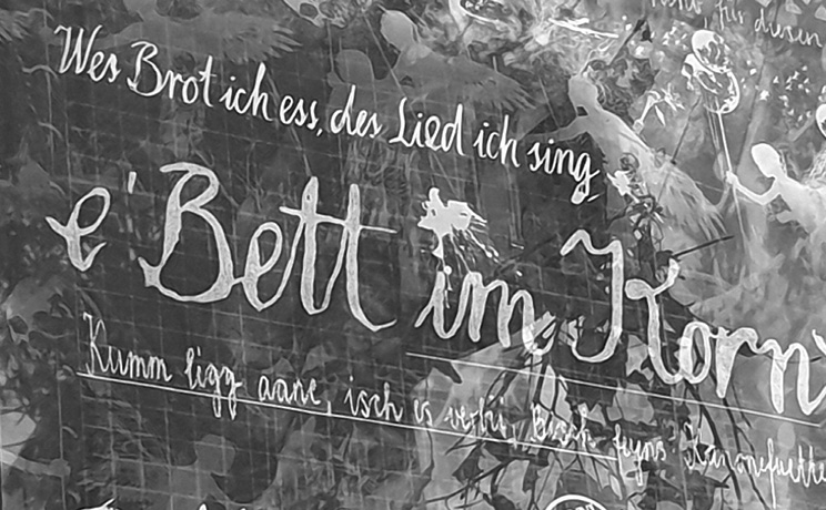 A wall with handwritten German text in white paint surrounded by faint images of dancing figures and foliage.
Description generated by AI
